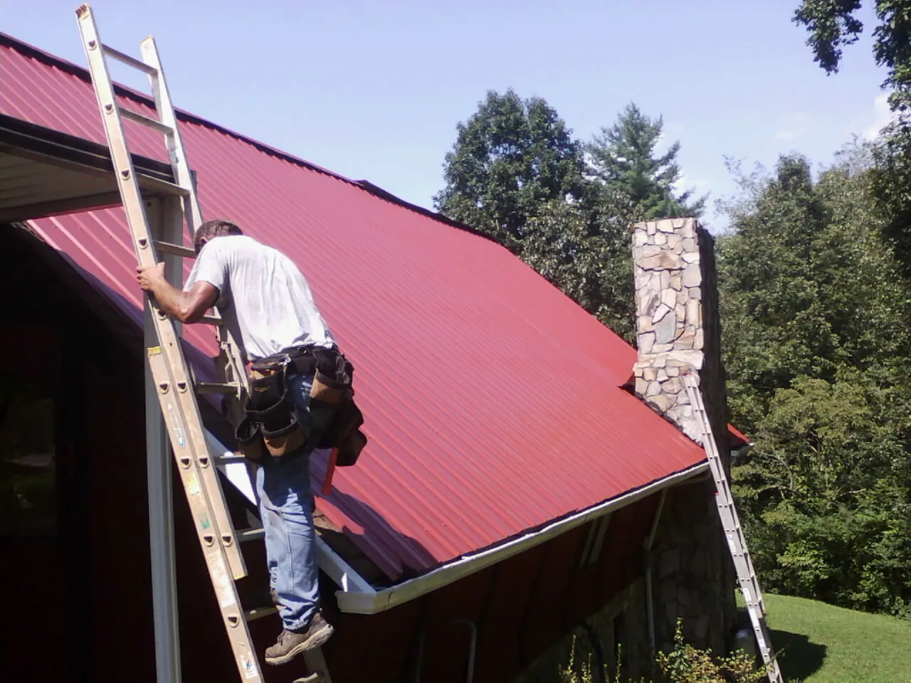 Roofer on a ladder beside a red metal roof and stone chimney — Soffit & Fascia Repair by Roofing Brothers in Rosemont